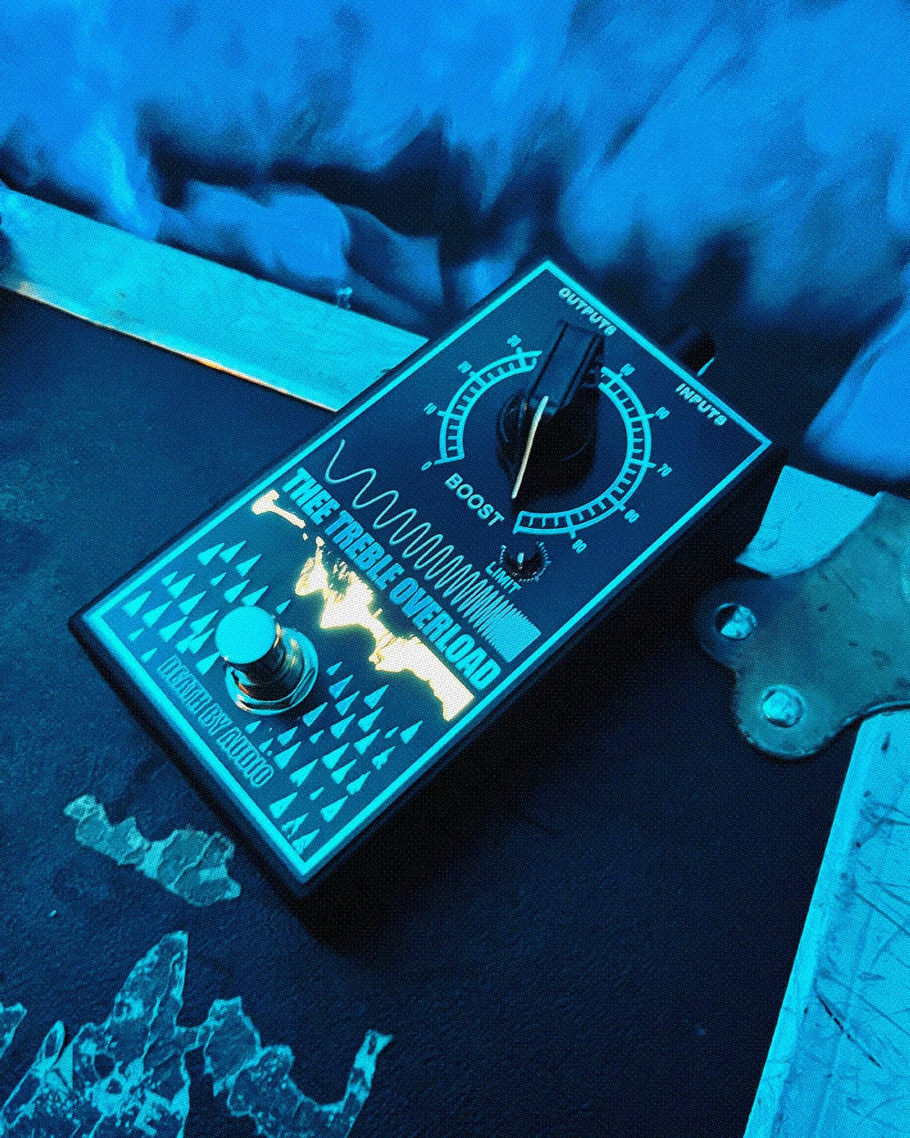 Death by Audio Thee Treble Overload photographed on a weathered plank under blue stage lighting and haze