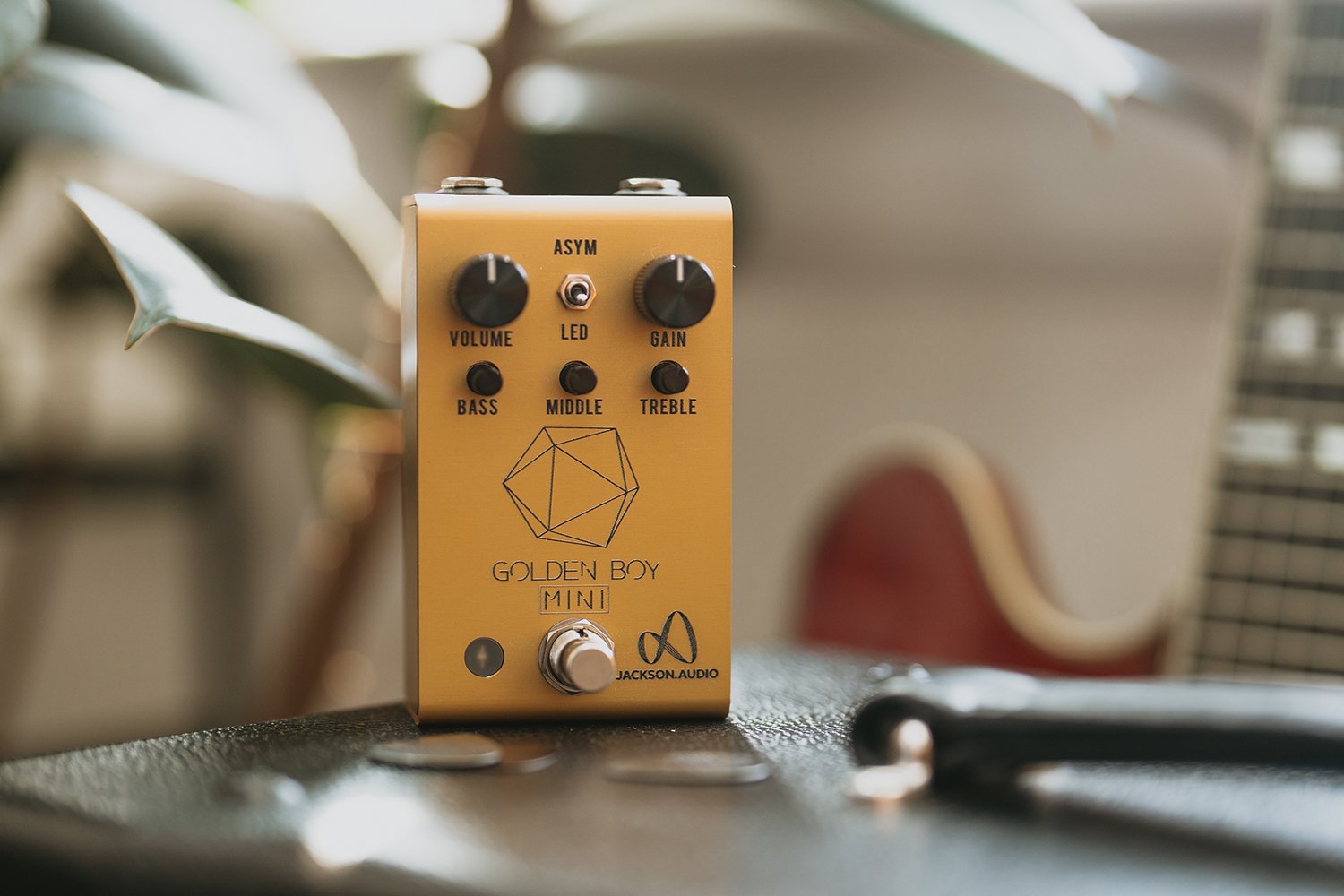 Jackson Audio Golden Boy Mini Transparent Overdrive, Lifestyle photo of the Golden Boy Mini positioned near a guitar case.