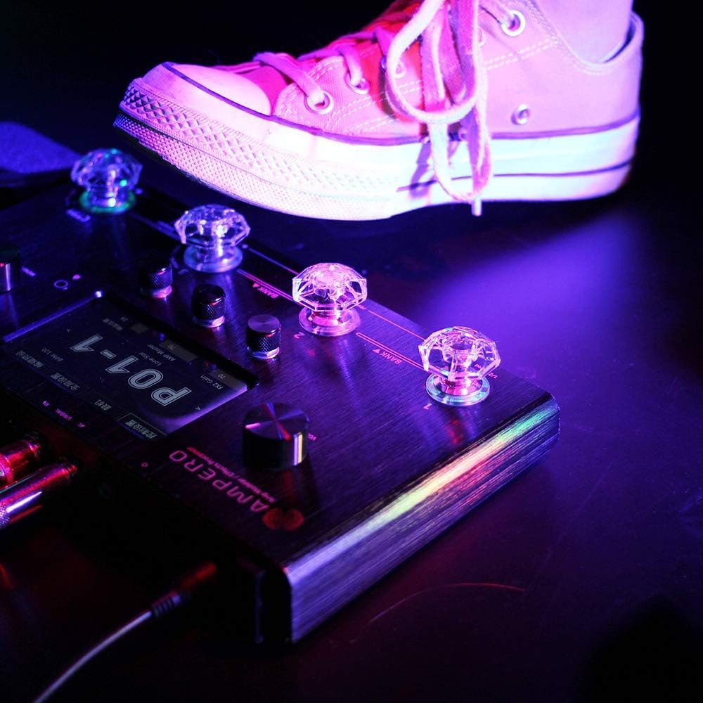 Musician's sneaker beside a pedalboard fitted with glowing Hotone Karat Cap footswitch covers, stage lighting