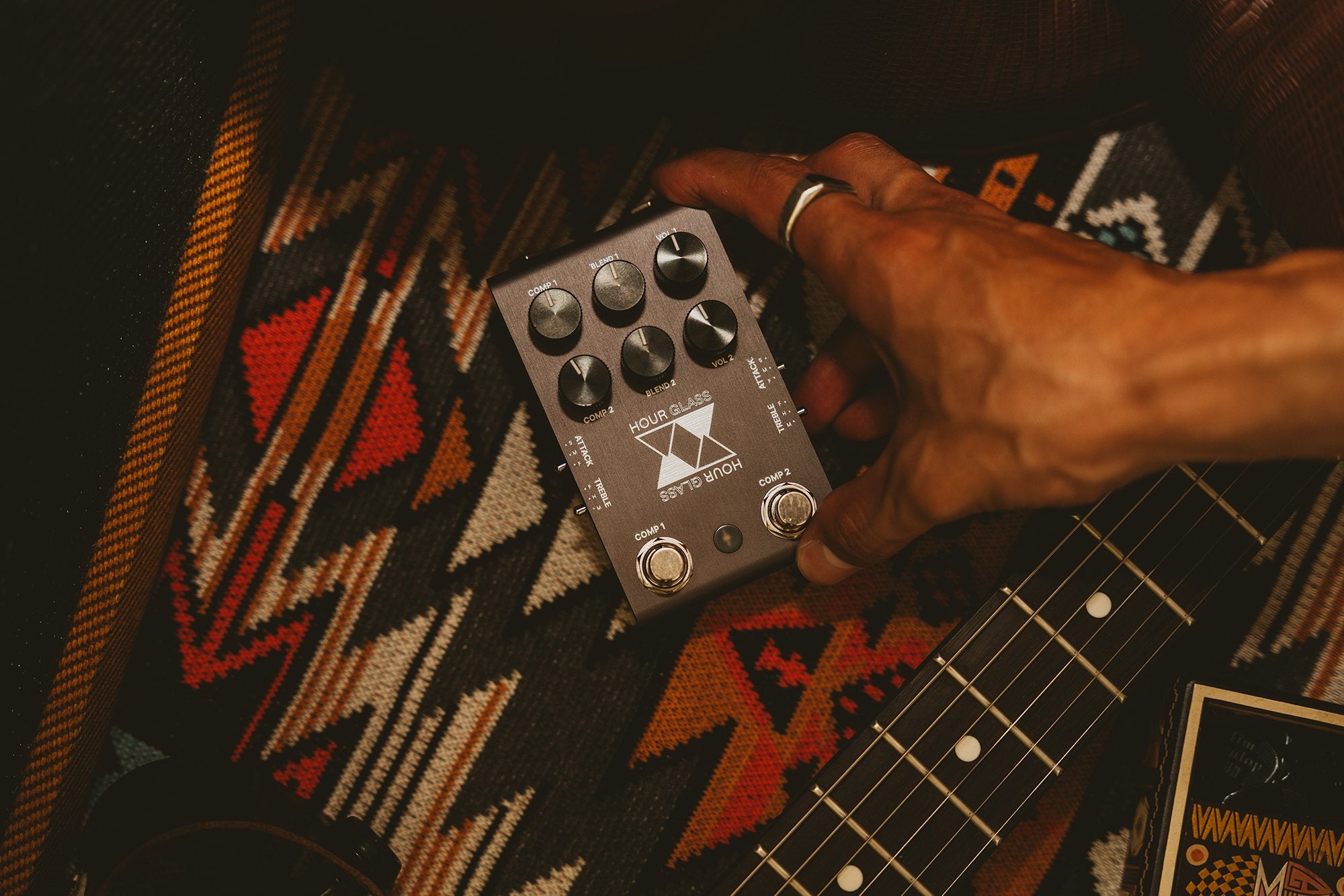 Jackson Audio Hour Glass Analog Dual Compressor Pedal, Lifestyle photo of a hand holding the Hour Glass pedal over a guitar and a patterned rug.
