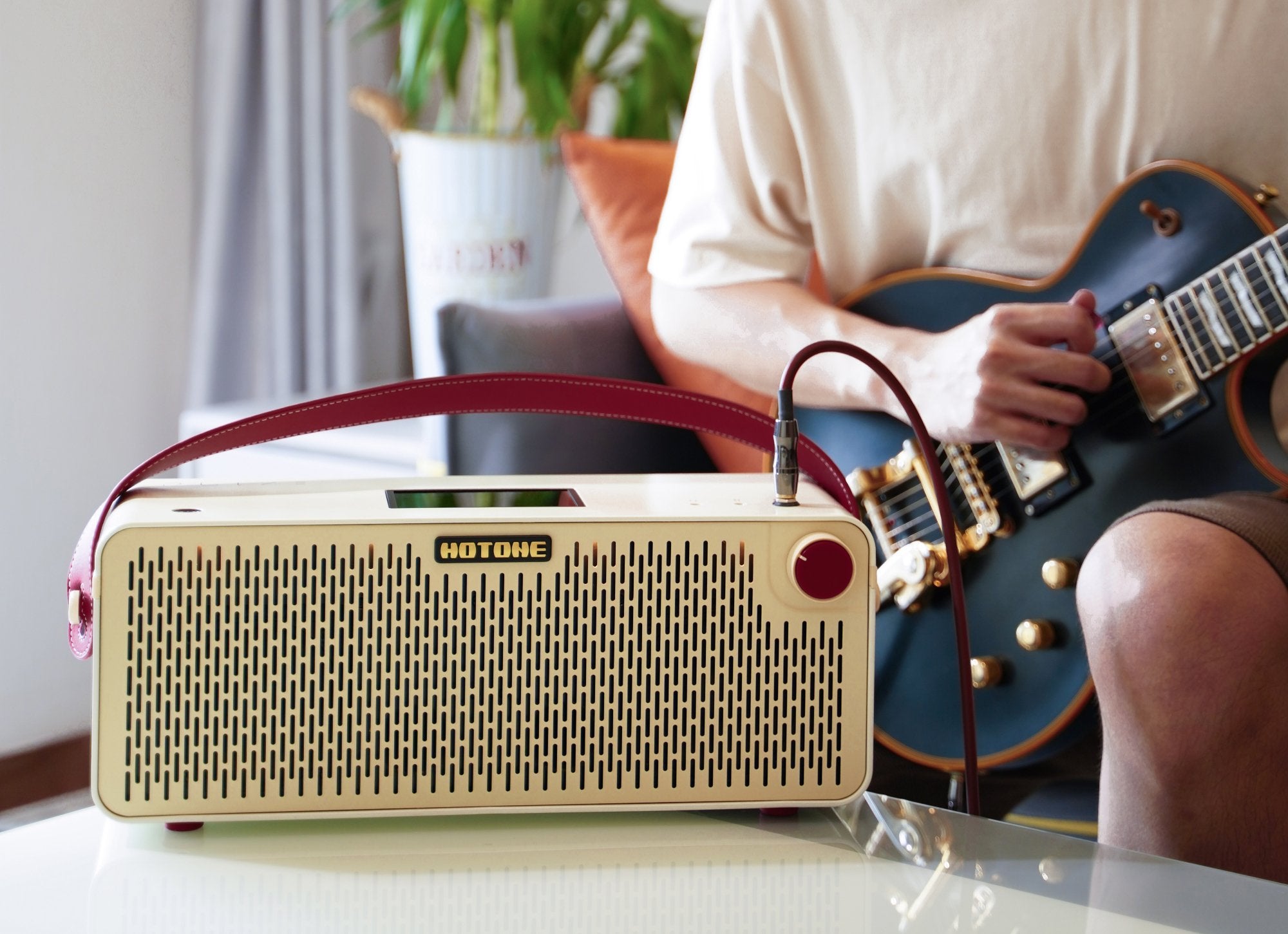 Person playing guitar on a couch beside a Hotone Pulze amplifier and houseplant